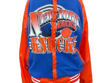 Könnte beinhalten: Blaue und orangefarbene Satinjacke mit der Aufschrift "NEW YORK KNICKS" und einer Basketballgrafik. Die Jacke hat eine Knopfleiste, weiß-orange gestreifte Bündchen und einen weiß-orange gestreiften Kragen.