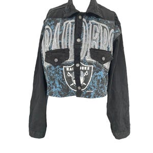 May include: Black cropped denim jacket with long sleeves. The jacket features a Raiders logo and text on the front, with a blue and white marbled pattern. It has two front pockets and button closures.