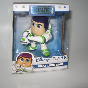 Könnte beinhalten: Eine Plastikfigur von Buzz Lightyear aus dem Disney Pixar Film Toy Story. Die Figur befindet sich in einer durchsichtigen Plastikbox mit weißem Hintergrund und dem Aufdruck "Disney Pixar Buzz Lightyear" auf der Vorderseite. Die Figur trägt einen weißen und grünen Raumanzug mit einem violetten Helm und hält eine grüne Laserpistole in der rechten Hand.
