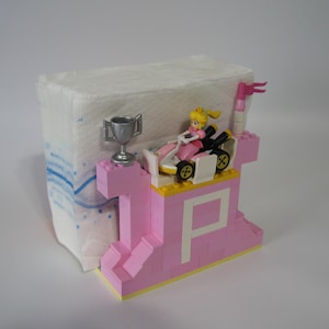 May include: A pink Lego castle with a white 'P' on the front. A small figure of Princess Peach is driving a kart with a silver trophy on top of the castle.
