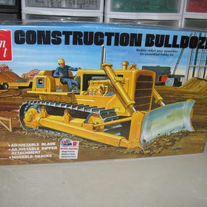 May include: A vintage model kit box for a construction bulldozer. The box art features a yellow bulldozer with a blue blade, a construction worker, and the text "CONSTRUCTION BULLDOZER". It includes features like an adjustable blade and movable tracks.