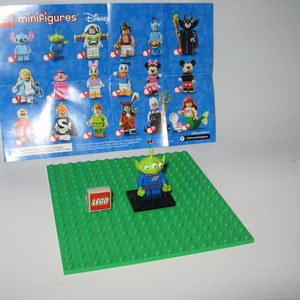 May include: A green alien Lego minifigure from the Toy Story movie series. The minifigure is standing on a black base plate. The minifigure is wearing a blue spacesuit with a green shirt underneath. The minifigure has a green head with three eyes and a big smile. The minifigure is holding a small black gun in its right hand. The minifigure is standing on a green Lego base plate. The minifigure is surrounded by a white background. The image also shows a small Lego box with the word "LEGO" printed on it.