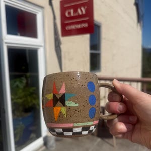 May include: A speckled tan ceramic mug featuring a multi-coloured star design, three blue circles, and a black and white chequered pattern along the base. The mug has a curved handle and a smooth, glazed finish with visible dark brown speckles.