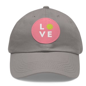 May include: A grey baseball cap with a pink circle patch that says "LOVE" with a yellow and green pickleball inside the circle.