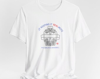 May include: White t-shirt with a gray catcher's mask graphic and the text "JT Keeping It Realmuto" in blue and red. The text "Philadelphia Baseball" and the number "18" are also printed on the shirt.