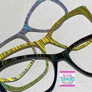 May include: Three pairs of eyeglass frames in silver, gold, and black, each with a striped, iridescent design. The frames are displayed on a white surface with glitter. The text "Top It Off Toppers" is visible.