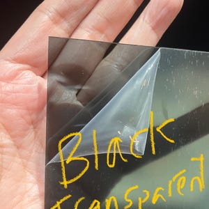 May include: A black, transparent sheet of material with a protective film being peeled back. The words "Black Transparent" are written in yellow marker on the sheet.
