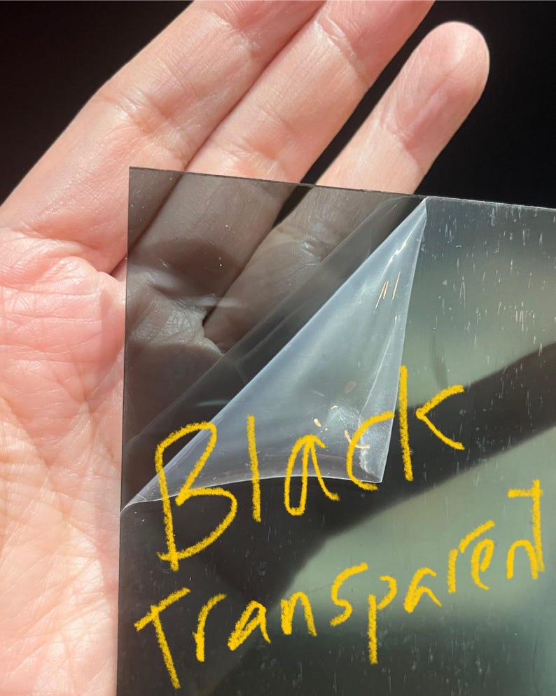 May include: A black, transparent sheet of material with a protective film being peeled back. The words "Black Transparent" are written in yellow marker on the sheet.