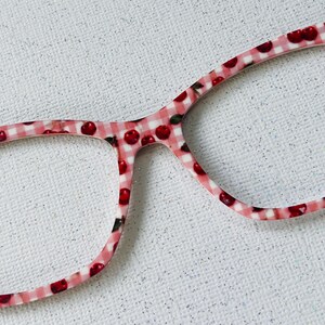 May include: Eyeglass frames featuring a pink and white gingham check pattern with a repeating print of small red cherries. The frames have a smooth, glossy finish and a classic rectangular shape with rounded edges, showcasing a vibrant design.
