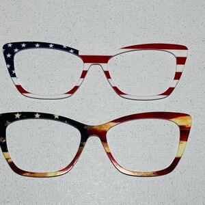 May include: Two pairs of eyeglasses with patriotic designs. One pair features a red, white, and blue American flag pattern. The other pair has a similar design with a sunset-like gradient. Both pairs have a cat-eye shape.