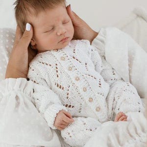 May include: A sleeping baby in a white, open-knit romper with wooden buttons. The baby is cradled in someone's arms, with hands gently supporting the head. The romper features a delicate, lacy pattern.