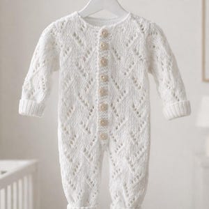 Può includere: Un pagliaccetto per neonati bianco lavorato a maglia con un delicato motivo traforato. Il pagliaccetto a maniche lunghe presenta un'abbottonatura frontale e polsini e caviglie a coste. L'indumento è appeso a un'appendiabiti bianco.