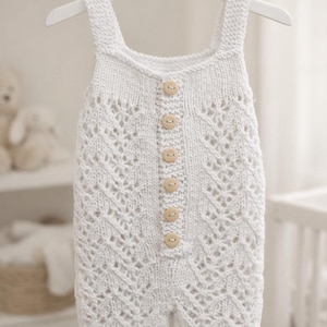 Könnte beinhalten: Ein weißer gestrickter Baby-Strampler mit einem zarten Lochmuster und Holzknöpfen auf der Vorderseite. Der Strampler hängt an einem weißen Kleiderbügel und zeigt sein aufwendiges Design und seine weiche Textur. Ein charmantes und gemütliches Kleidungsstück.