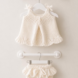 Puede incluir: Un conjunto de dos piezas tejido en color crema que consta de una parte superior sin mangas con tirantes para anudar y un cuerpo texturizado, junto con bombachos a juego con cintura con volantes, cordón decorativo y patrón de punto.