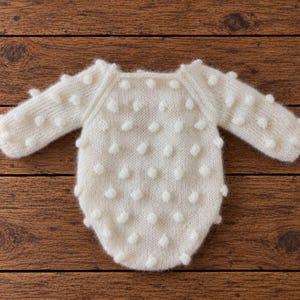 May include: A long-sleeved, cream-colored knit romper featuring a textured bobble pattern across the front and sleeves. The garment has a rounded neckline and a soft, fuzzy appearance, showcasing a consistent knit construction throughout.