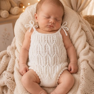 Può includere: Una tutina lavorata a maglia di colore bianco con un delicato motivo in pizzo a forma di foglia, spalline sottili con piccoli fiocchi e un bordo smerlato lungo le aperture delle gambe. Il capo è realizzato in filato morbido per neonati.