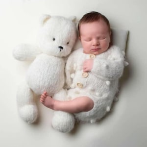 Può includere: Un neonato addormentato, vestito con una tutina bianca lavorata a maglia con bottoni, accanto a un orsacchiotto bianco. Il bambino riposa su un piccolo cuscino. L'orsacchiotto ha naso e occhi neri.