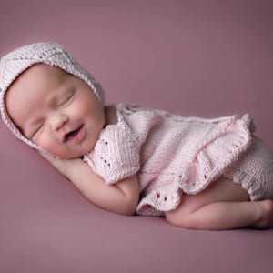 May include: A newborn baby wearing a light pink knitted outfit and bonnet, lying on a pink surface. The outfit has a ruffled trim and the baby is smiling with eyes closed.