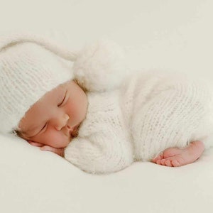 May include: A newborn baby sleeping soundly in a white knitted hat and matching outfit. The baby is lying on a white background.