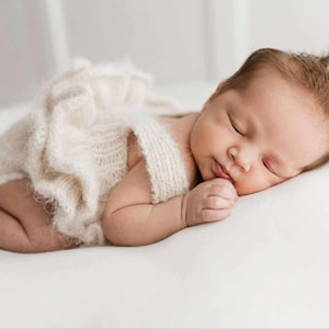 May include: A newborn baby sleeping peacefully on a white blanket, wearing a white crocheted outfit with ruffles.
