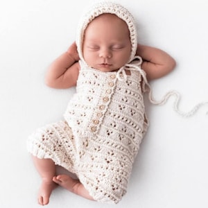 May include: A newborn baby wearing a cream-colored knitted romper and bonnet. The romper has a button-up front and decorative openwork. The baby is lying on its back with its hands behind its head, eyes closed.