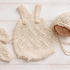 Pode incluir: Um conjunto de roupa de bebé de malha creme, incluindo um macacão com alças, um gorro e duas luvas a condizer. O macacão tem um padrão decorativo vazado e botões de madeira. O conjunto é exibido numa superfície de madeira branca.