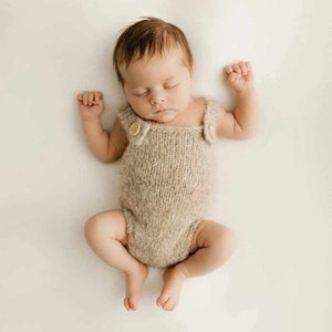 May include: A sleeping baby wearing a beige knitted onesie with wooden buttons. The baby is lying on its back with its arms outstretched and its legs crossed.