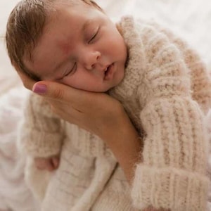 May include: A newborn infant with light brown hair is sleeping peacefully while wearing a cream-colored, long-sleeved, chunky knit sweater. The sweater features a ribbed texture on the sleeves and a soft, fuzzy appearance throughout the garment.