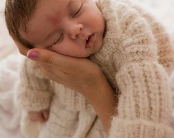 May include: A newborn infant with light brown hair is sleeping peacefully while wearing a cream-colored, long-sleeved, chunky knit sweater. The sweater features a ribbed texture on the sleeves and a soft, fuzzy appearance throughout the garment.