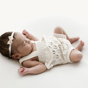 May include: A sleeping newborn baby wearing a cream-coloured knitted romper and a matching headband with a bow. The baby is lying on a white surface. The knitted romper is made of a soft, open weave material.