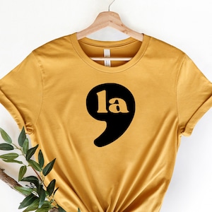 May include: A mustard yellow t-shirt with a black semicolon graphic and the word "la" inside the semicolon.