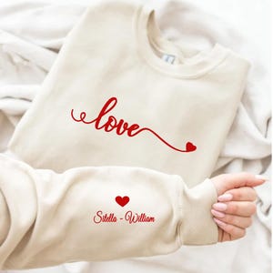 May include: A cream-colored sweatshirt with the word "love" in red script across the chest, with a heart at the end of the word. The sleeve features a red heart and the names "Sitella - William".