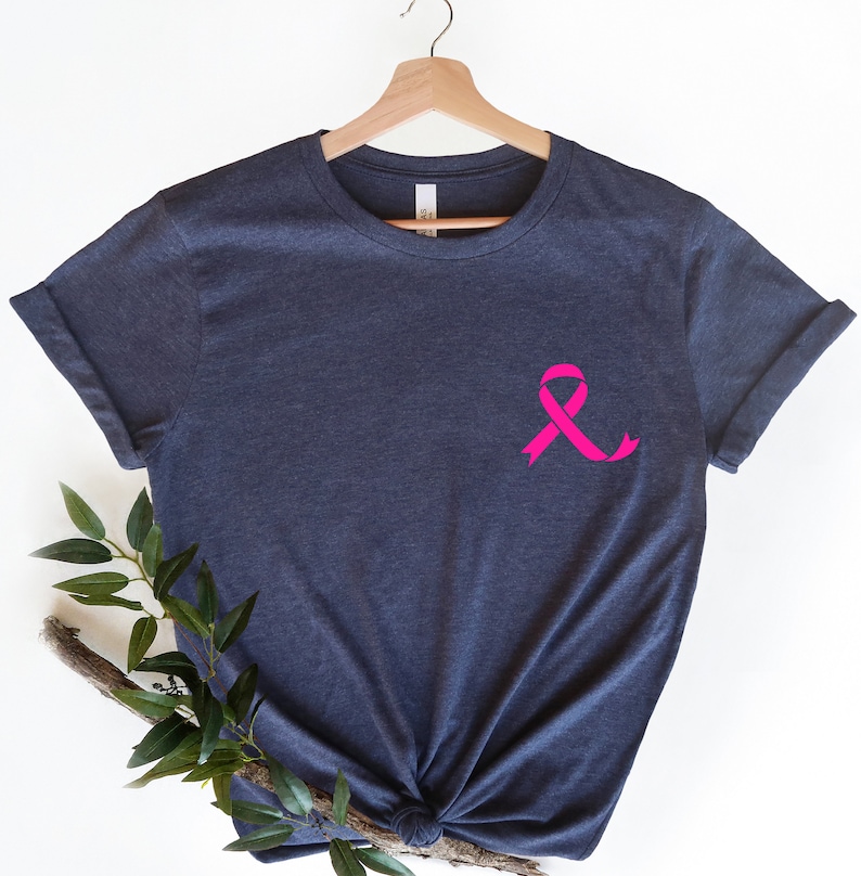 K&ouml;nnte beinhalten: Ein dunkelblaues T-Shirt mit einem rosa Band auf der Vorderseite. Das Band ist ein Symbol f&uuml;r die Sensibilisierung f&uuml;r Brustkrebs.