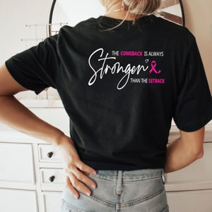 K&ouml;nnte beinhalten: Schwarzes T-Shirt mit wei&szlig;em Text, der "The comeback is always stronger than the setback" lautet, mit einem rosa Band und einem Herzsymbol.
