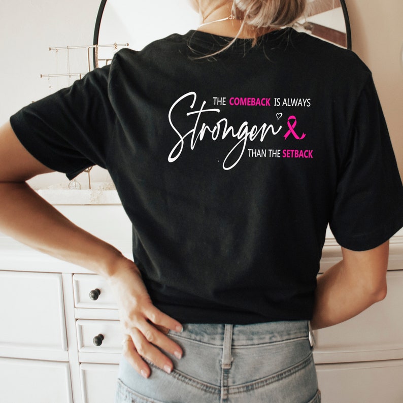 K&ouml;nnte beinhalten: Schwarzes T-Shirt mit wei&szlig;em Text, der "The comeback is always stronger than the setback" lautet, mit einem rosa Band und einem Herzsymbol.