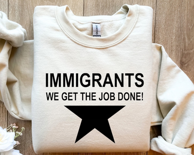 May include: A beige sweatshirt with the text "Immigrants We Get The Job Done!" and a black star printed on the front.