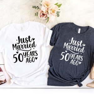 May include: Two t-shirts with the text "Just Married 50 Years Ago" printed on them. The shirts are white and dark grey. The text is in black on the white shirt and white on the dark grey shirt.