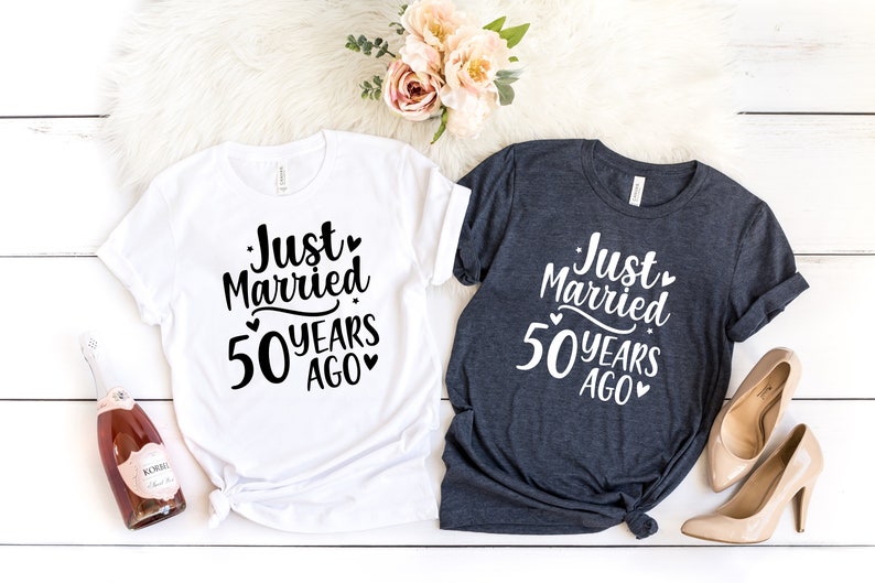 May include: Two t-shirts with the text "Just Married 50 Years Ago" printed on them. The shirts are white and dark grey. The text is in black on the white shirt and white on the dark grey shirt.