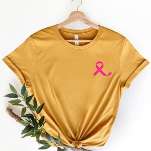 K&ouml;nnte beinhalten: Ein senfgelbes T-Shirt mit einem rosa Band auf der linken Brust. Das Band ist ein Symbol f&uuml;r die Sensibilisierung f&uuml;r Brustkrebs.