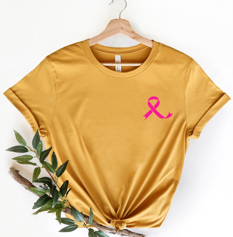 K&ouml;nnte beinhalten: Ein senfgelbes T-Shirt mit einem rosa Band auf der linken Brust. Das Band ist ein Symbol f&uuml;r die Sensibilisierung f&uuml;r Brustkrebs.