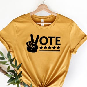 May include: A mustard yellow short-sleeved t-shirt featuring a black graphic print on the chest. The design includes a hand making a peace sign, the word VOTE in bold capital letters, five black stars, and two horizontal lines underneath.