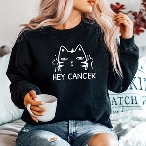 May include: Black sweatshirt with a white graphic of a cat giving the middle finger with the text "Hey Cancer".