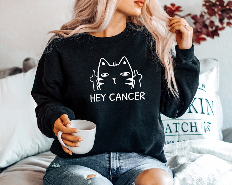May include: Black sweatshirt with a white graphic of a cat giving the middle finger with the text "Hey Cancer".