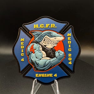 May include: A blue and black emblem featuring a muscular shark character with the number 4. The emblem has the text "H.C.F.R.", "MEDIC 4", "MEDIC 204", and "ENGINE 4" in yellow. The shark is flexing its muscles.