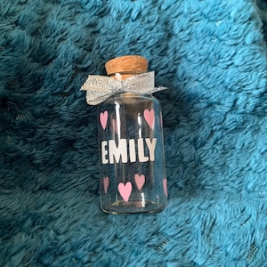 May include: A small glass bottle with a cork top and a silver ribbon bow. The bottle is decorated with pink hearts and the name "EMILY" in white lettering.