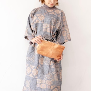 以下が含まれることがあります： グレーの柄のワンピースを着た女性が、お揃いのベルトとライトブラウンのクラッチバッグを持っています。ワンピースはグレーとベージュの色合いの幾何学模様です。クラッチバッグは長方形で、革のようです。