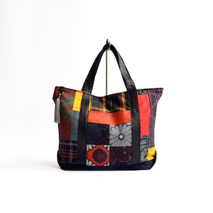 May include: A patchwork tote bag with a variety of colorful fabric squares, including plaid and floral patterns. The bag has black leather handles and accents, a black base, and a gray tassel zipper pull. The bag is displayed against a white background.