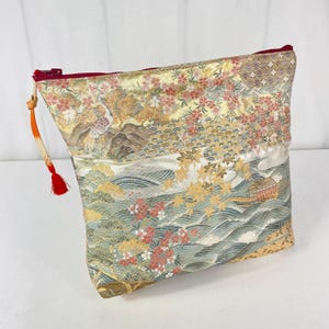 Può includere: Una pochette decorativa con cerniera e nappa rosse. La pochette presenta un design paesaggistico dettagliato con un pavone, montagne ed elementi floreali nei toni dell'oro, del verde e del rosa. La pochette è rettangolare e ha un interno rosso.