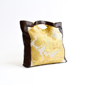 May include: A tote bag featuring a white fabric body with a repeating yellow and gold fan-shaped pattern. The bag has dark brown leather-like side panels and integrated handles at the top, with a single button closure visible at the center opening.