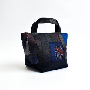 Puede incluir: Un bolso tote con diseño de patchwork en telas azul oscuro y negro con motivos florales. El bolso incluye asas con textura de cocodrilo en negro, una base negra sólida y una borla gris sujeta a una anilla plateada en el lateral.